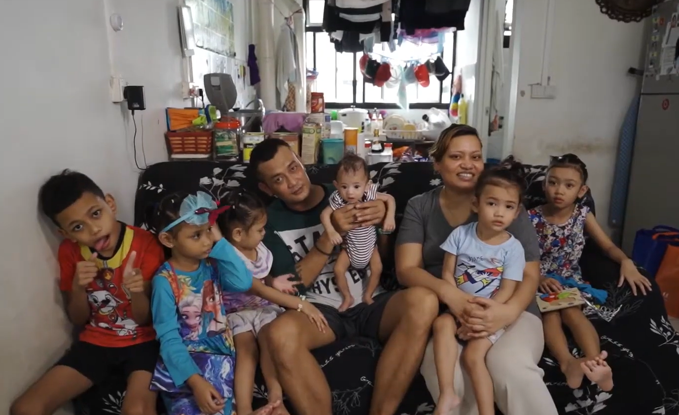 Family portrait taken at home, showing parents and several young children seated together in a modest living room setting.